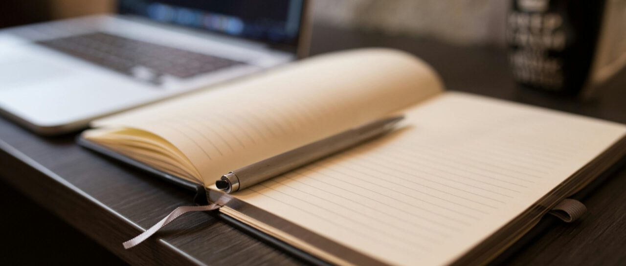 Bild eines Schreibtisches mit aufgeschlagenem Notizbuch, Laptop und einem Kaffeebacher